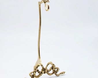 May include: A gold metal stand with a bow design at the top and bottom. The stand is designed to hold a small object, such as a necklace or bracelet.