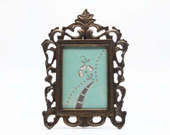 May include: Ornate bronze-colored picture frame with a rectangular opening. The frame has a detailed, scrolling design. The picture inside is a light blue with a floral design and a dotted border.