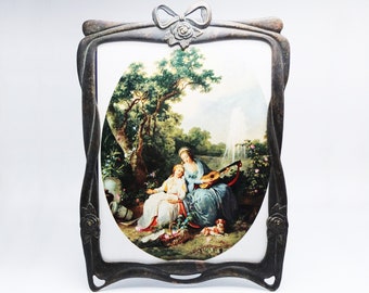 May include: An ornate black metal picture frame with a bow at the top. The frame holds a vintage oval-shaped print of two women in a garden setting, one playing a lute.