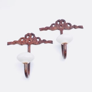 May include: Two wall hooks featuring a dark, textured, rust-colored metal backplate with an ornate, ribbon-like design. Each hook has a smooth, white, rounded ceramic knob attached to a curved metal arm extending from the center of the backplate.