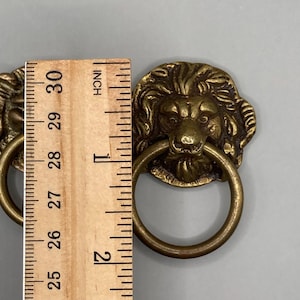 Puede incluir: Dos tiradores de cajones de cabeza de le&oacute;n de lat&oacute;n antiguo con asas de anillo. Cada tirador mide aproximadamente 2,5 cm de di&aacute;metro.