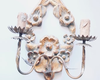 May include: A wall sconce featuring a carved wooden backplate with floral motifs in a distressed gold finish. Two metal arms extend outward, each supporting a candle-style light fixture with a serrated metal drip pan and a cylindrical base.