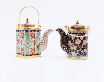 May include: Two miniature teapots with intricate floral designs. One teapot is white with blue, green, and red flowers, while the other is black with white and blue flowers. Both teapots have gold accents.