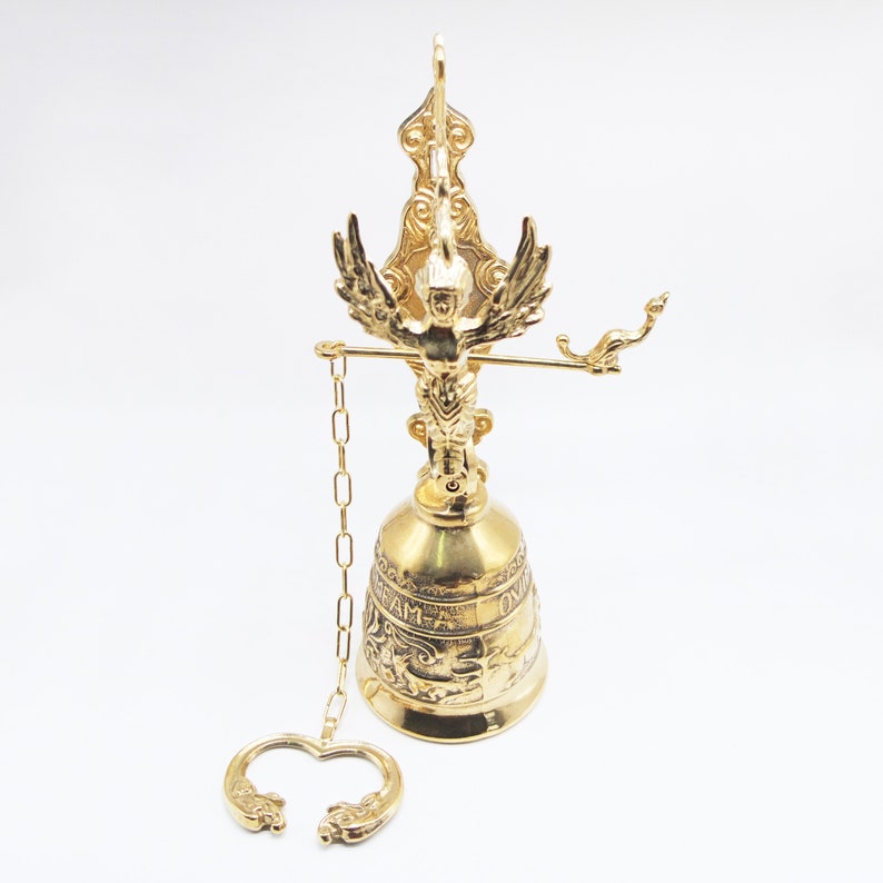 Op de afbeelding: Een gouden metalen bel met een sierlijk ontwerp. De bel is bevestigd aan een ketting en heeft bovenop een decoratief adelaarfiguur. De bel heeft de inscripties "Ave Maria" en "Ora Pro Nobis".