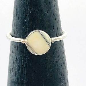 Puede incluir: Un anillo de plata con una incrustación redonda blanca y gris. El anillo está en un soporte de anillo de madera.