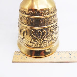 Op de afbeelding: Een gouden bel met een sierlijk ontwerp en de inscriptie "OCEM ME AME" aan de zijkant. De bel is ongeveer 18 cm hoog en staat op een houten liniaal.