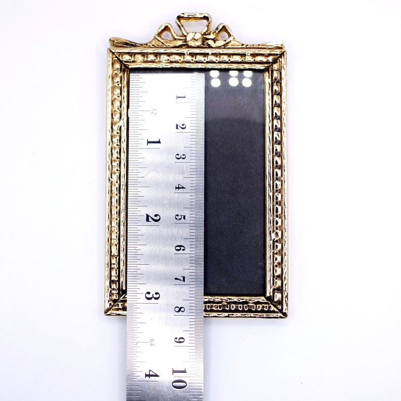 May include: A gold-colored picture frame with a decorative design around the edge. The frame is 4 inches tall and 3 inches wide.
