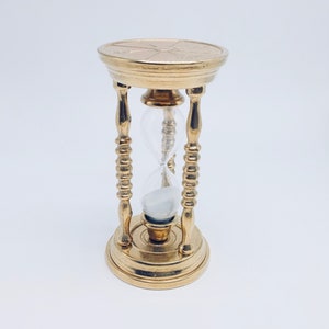 May include: A gold hourglass with a decorative base and a clear glass bulb. The hourglass is filled with white sand.