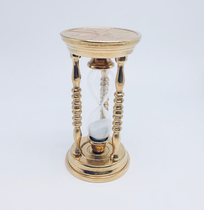 May include: A gold hourglass with a decorative base and a clear glass bulb. The hourglass is filled with white sand.