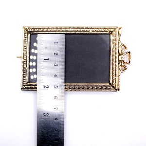 May include: A gold-colored picture frame with a black mat and a ruler measuring 7 inches in length. The frame has a decorative design around the edge and a small loop on the top for hanging.