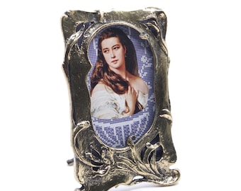 Puede incluir: Un marco de fotos de metal dorado con un diseño ornamentado. El marco presenta un retrato de una mujer con cabello largo y castaño. El marco tiene un soporte en la parte posterior.