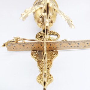 Op de afbeelding: Een gouden metalen wandklok met sierlijke details. De klok is bevestigd aan een decoratieve beugel met een rolontwerp. De klok is ongeveer 18 cm lang.