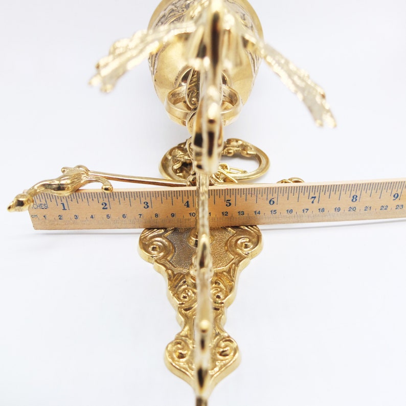Op de afbeelding: Een gouden metalen wandklok met sierlijke details. De klok is bevestigd aan een decoratieve beugel met een rolontwerp. De klok is ongeveer 18 cm lang.
