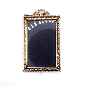 May include: A gold-colored picture frame with a decorative bow at the top. The frame is empty and has a black background.