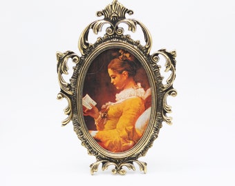 Puede incluir: Un marco de fotos adornado en tono dorado con un diseño detallado. El marco presenta una abertura ovalada con una imagen vintage de una mujer leyendo un libro.