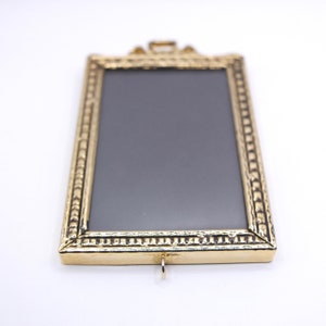 May include: A gold-toned picture frame with a textured border and a black background. The frame has a small loop at the top for hanging.