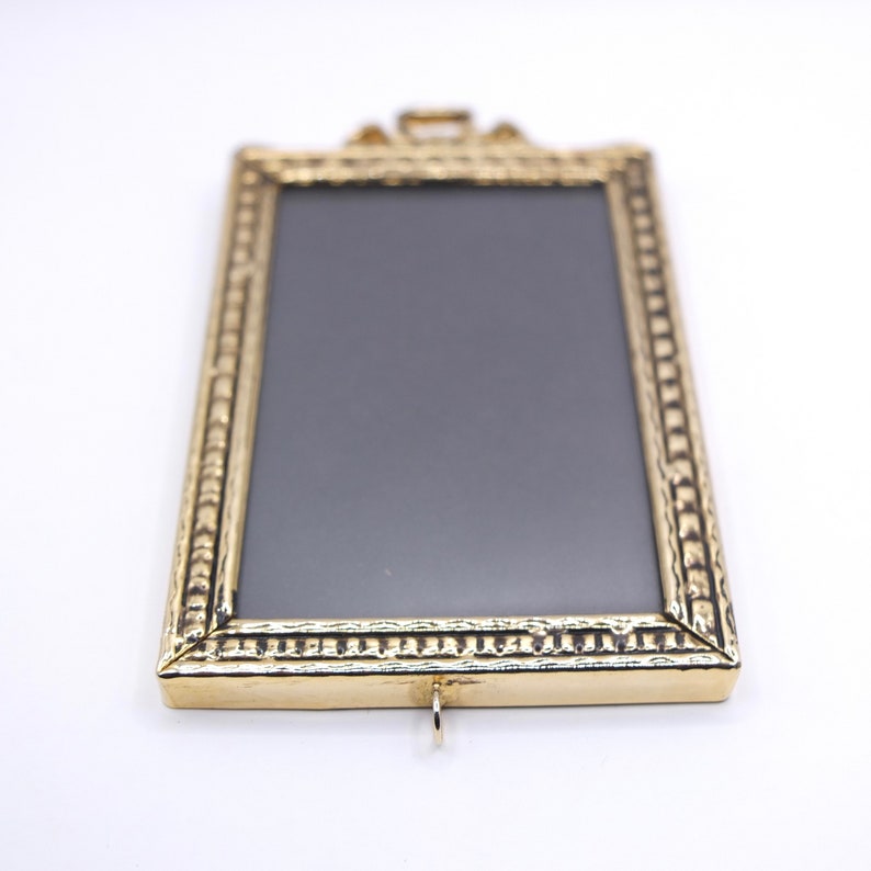 May include: A gold-toned picture frame with a textured border and a black background. The frame has a small loop at the top for hanging.