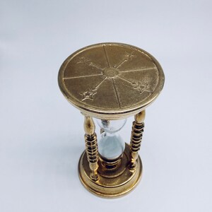 May include: A gold hourglass with a decorative top featuring a sunburst design and four stars. The hourglass is filled with sand and has a clear glass bulb.