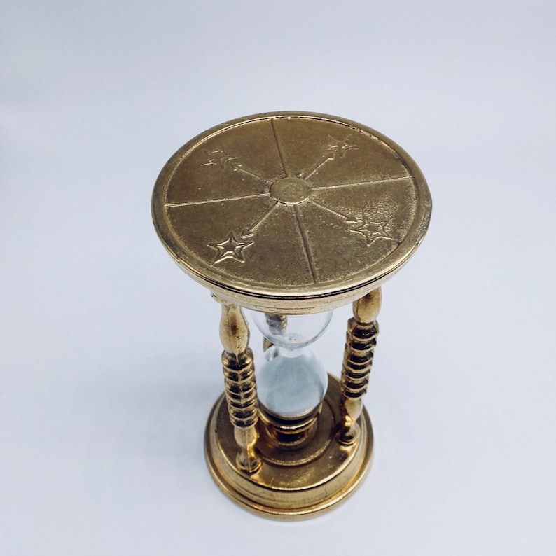 May include: A gold hourglass with a decorative top featuring a sunburst design and four stars. The hourglass is filled with sand and has a clear glass bulb.