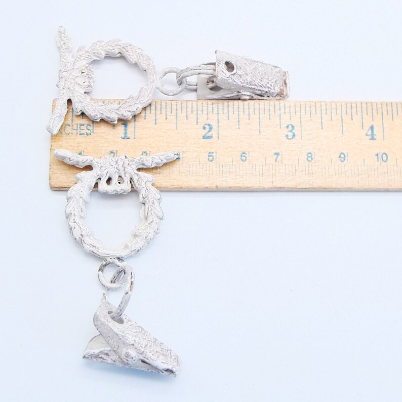 May include: Two silver-toned metal curtain tiebacks with a wreath design. The tiebacks are approximately 5 cm long and are shown next to a ruler.
