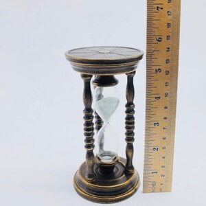 May include: A brass hourglass with a decorative base and a clear glass bulb. The hourglass is 6 inches tall.
