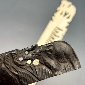 May include: A dark brown carved wooden letter opener featuring an elephant motif with small inlaid circular accents, positioned in front of a second, lighter-colored, off-white carved letter opener with a similar textured elephant design.
