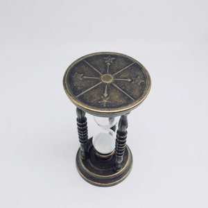 May include: A brass hourglass with a decorative top featuring a compass design. The hourglass is filled with sand and has a clear glass bulb.