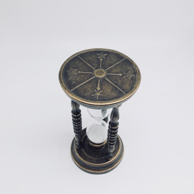 May include: A brass hourglass with a decorative top featuring a compass design. The hourglass is filled with sand and has a clear glass bulb.