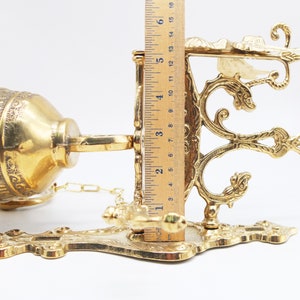 Op de afbeelding: Een gouden metalen wandklok met een sierlijk ontwerp. De klok is bevestigd aan een ketting en heeft een decoratieve haak.