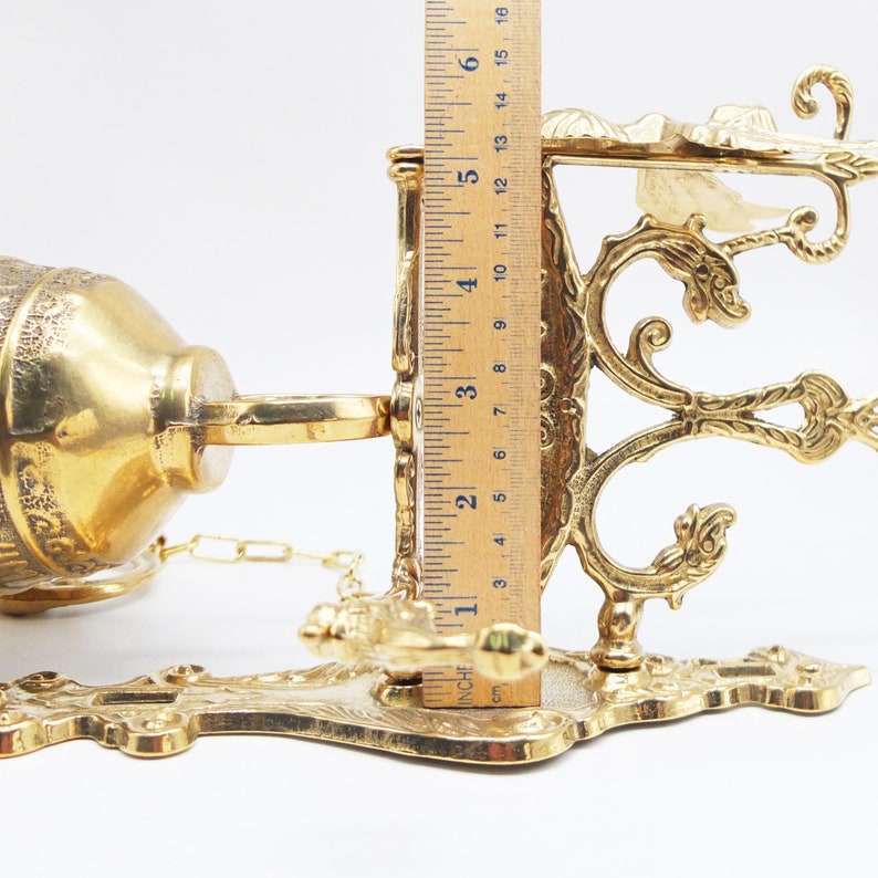Op de afbeelding: Een gouden metalen wandklok met een sierlijk ontwerp. De klok is bevestigd aan een ketting en heeft een decoratieve haak.