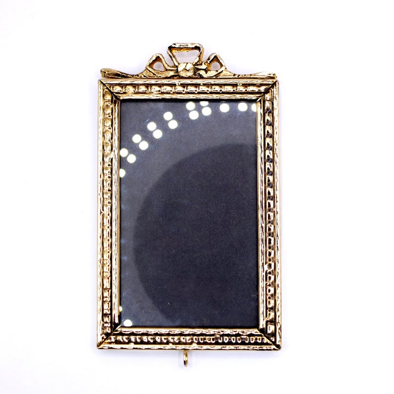 May include: A gold-colored picture frame with a decorative ornate design. The frame has a clear glass front and a black backing.
