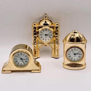 May include: Three gold-colored miniature clocks. One clock has an arched design, another is dome-shaped, and the third is a rectangular shape. Each clock has a white clock face with black Roman numerals and black hands.