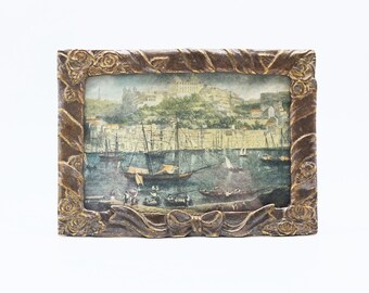 Puede incluir: Un marco de fotos de estilo vintage con un acabado dorado y detalles ornamentados. El marco presenta un lazo elevado en la parte inferior y una escena de un puerto con barcos y una ciudad en el fondo.