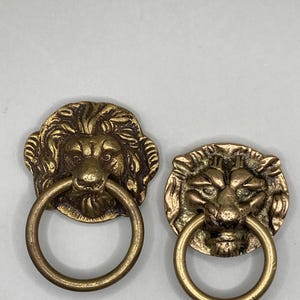 Puede incluir: Dos pomos de puerta antiguos de lat&oacute;n en forma de cabeza de le&oacute;n con tiradores de anillo. Las cabezas de le&oacute;n est&aacute;n detalladas con pelo texturizado y tienen una expresi&oacute;n realista.
