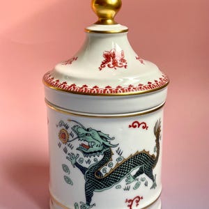 Pode incluir: Um pote de cerâmica branco com tampa e acabamento dourados. O pote apresenta uma ilustração detalhada de um dragão verde e preto segurando um sol. Desenhos florais vermelhos e nuvens decoram o pote.