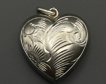 May include: A silver-toned heart-shaped pendant featuring an engraved floral pattern with swirling lines and textured details. The pendant has a smooth, polished finish and includes a small metal loop at the top for attachment to a chain or cord.