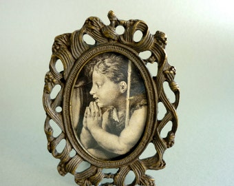 May include: A vintage oval-shaped brass picture frame with an ornate design. The frame features a black and white photo of a child praying.