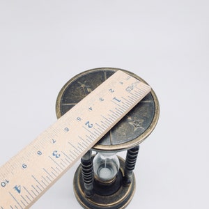 May include: A brass hourglass with a decorative top featuring a star and other symbols. The hourglass is 2 inches tall and is sitting on a white surface.
