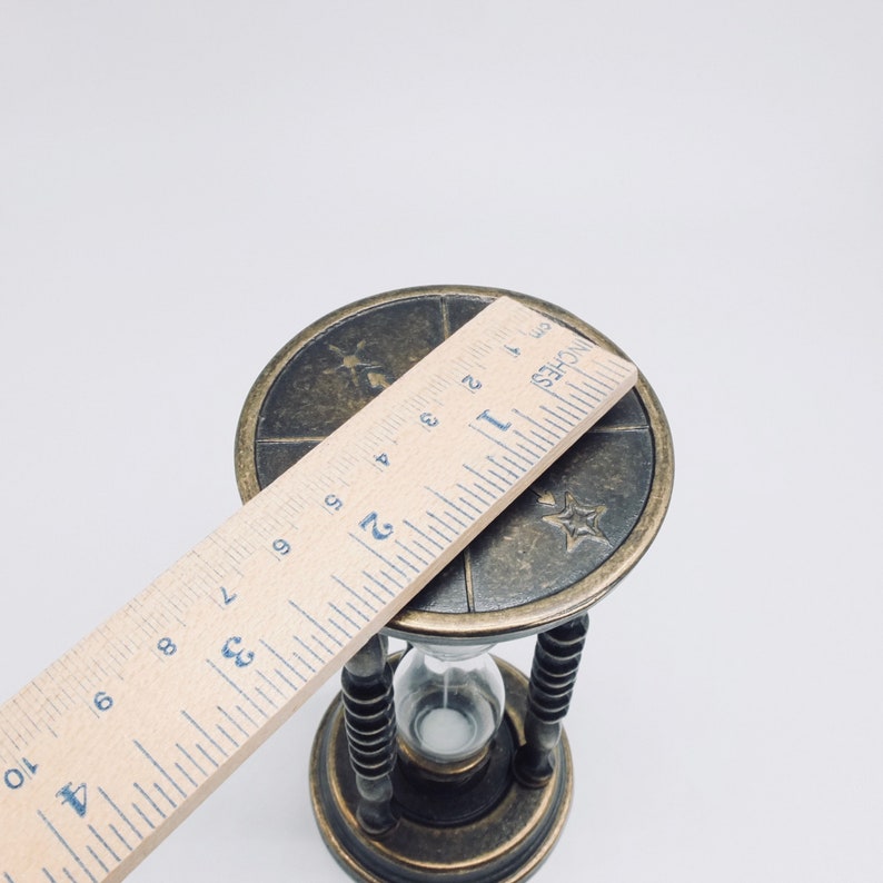 May include: A brass hourglass with a decorative top featuring a star and other symbols. The hourglass is 2 inches tall and is sitting on a white surface.