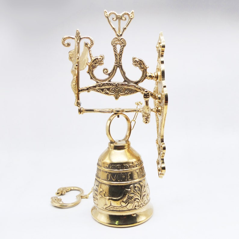 Op de afbeelding: Een gouden messing bel met een sierlijk ontwerp en een ophangbeugel. De bel heeft een decoratief opschrift met de tekst "OVME VA".