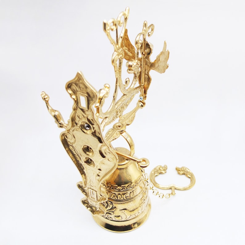 Op de afbeelding: Een gouden metalen bel met ingewikkelde details en een ketting. De bel heeft een decoratieve bovenkant met een vogelontwerp en het woord "Engel" is aan de zijkant gegraveerd.