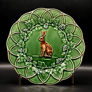 May include: A round green ceramic plate featuring a central embossed brown rabbit. The plate is decorated with a raised white grape vine and leaf pattern around the border, with a scalloped edge and a glossy finish throughout the surface.