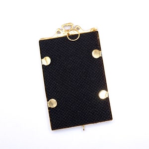 May include: A gold-colored picture frame with a black fabric backing and four gold-colored accents. The frame has a decorative top with a ring.