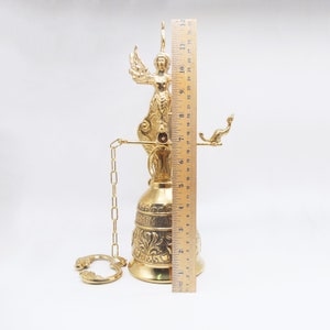 Op de afbeelding: Een gouden metalen bel met een engelfiguur erop. De bel hangt aan een ketting en heeft een decoratief ontwerp. De bel is 30,5 cm hoog.