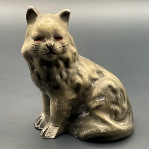 Peut inclure: Une figurine de chat en céramique grise en position assise avec une finition texturée et tachetée. Le chat a des yeux orange peints, des moustaches noires et un petit nez. Il y a un éclat visible sur la patte avant gauche et de l'usure.