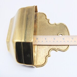 Puede incluir: Un macetero de pared de metal dorado con una abertura rectangular y un borde inferior decorativo y ornamentado. El macetero mide aproximadamente 18 cm de ancho.