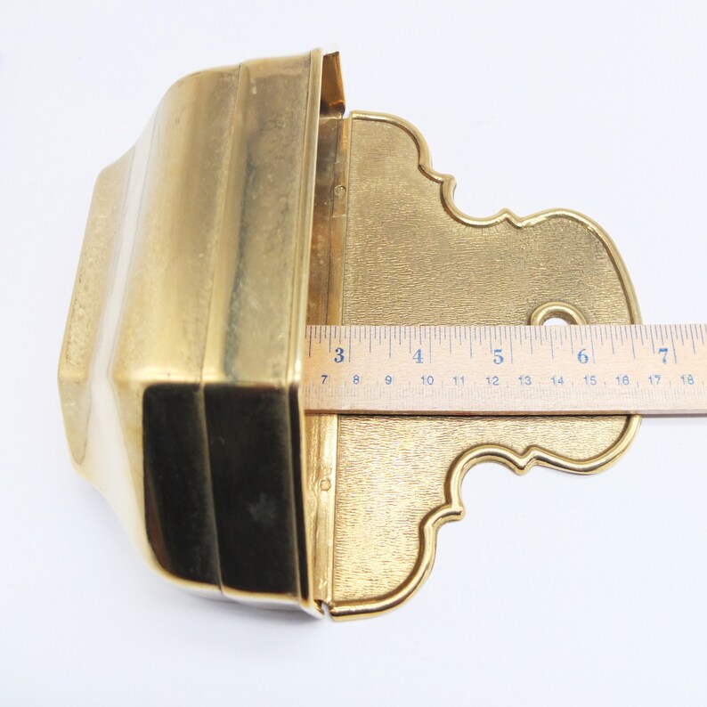 Puede incluir: Un macetero de pared de metal dorado con una abertura rectangular y un borde inferior decorativo y ornamentado. El macetero mide aproximadamente 18 cm de ancho.