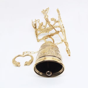 Op de afbeelding: Een gouden metalen bel met ingewikkelde bloemmotieven en een ketting. De bel is bevestigd aan een wandbevestiging met een decoratief ontwerp.
