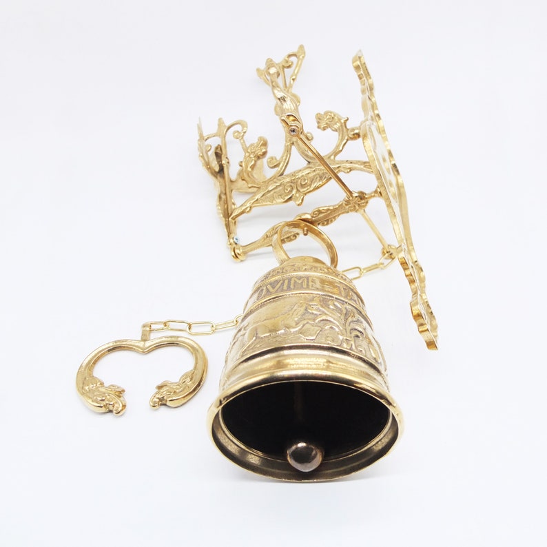 Op de afbeelding: Een gouden metalen bel met ingewikkelde bloemmotieven en een ketting. De bel is bevestigd aan een wandbevestiging met een decoratief ontwerp.
