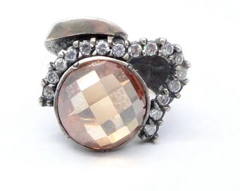 May include: A silver ring featuring a large, faceted, peach-colored gemstone. The gemstone is surrounded by a halo of small, clear crystals. The ring has a unique, artistic design with a heart-shaped element on one side.